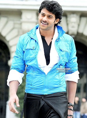 Prabhas in a bright blue jacket and black trousers, smiling outdoors in front of a stone archway HD phone wallpaper