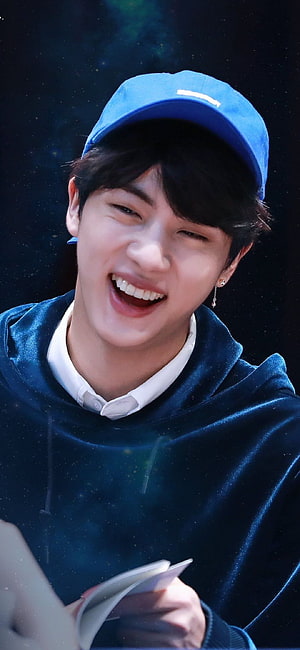 BTS Jin smiling in a blue cap and hoodie with a dark starry background HD phone wallpaper