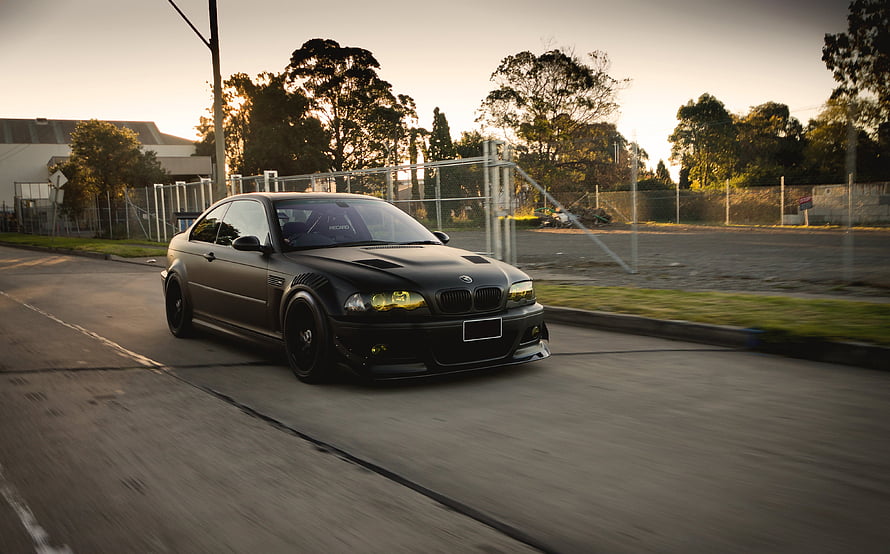 Black BMW E46 M3 Coupe with Widebody Kit Driving on a Suburban Road at Sunset HD wallpaper
