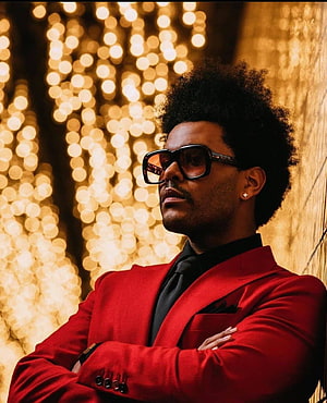 The Weeknd in a red suit with bold glasses against a golden bokeh background HD phone wallpaper
