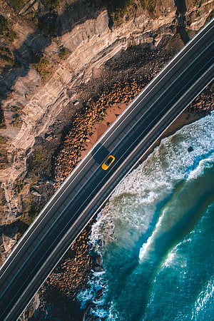 Aerial View of Yellow Car Driving on Coastal Highway Beside Turquoise Ocean Waves HD phone wallpaper