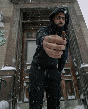 Bad Bunny in black hoodie and beanie holding a cigar in falling snow outside a brownstone HD phone wallpaper