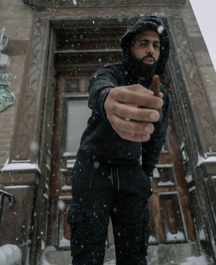 Bad Bunny in black hoodie and beanie holding a cigar in falling snow outside a brownstone HD phone wallpaper