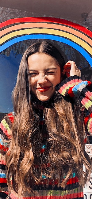 Olivia Rodrigo smiling in colorful striped sweater under a rainbow arch HD phone wallpaper