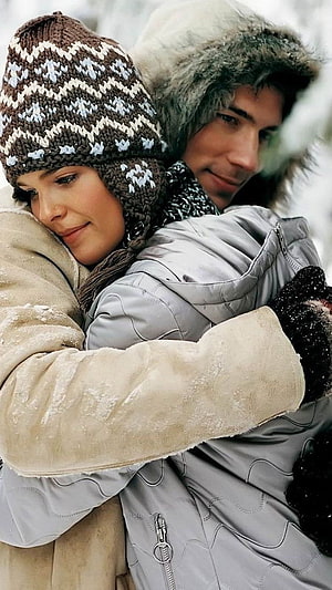 Couple embracing in winter coats and knit hats during a snowy romantic moment HD phone wallpaper
