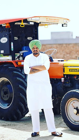 Sidhu Moose Wala in white kurta and green turban standing before a large orange tractor HD phone wallpaper