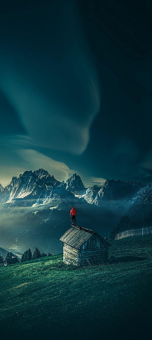 Lone Hiker on Alpine Cabin Roof with Dramatic Mountain Peaks and Misty Valley HD phone wallpaper