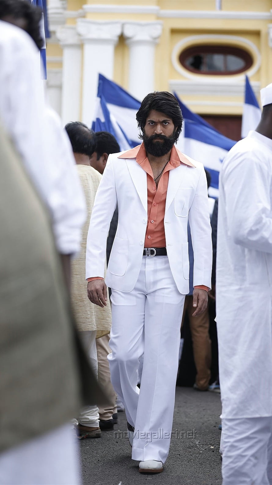 Yash in a white suit and salmon shirt walking through a crowd with blue and white flags HD phone wallpaper