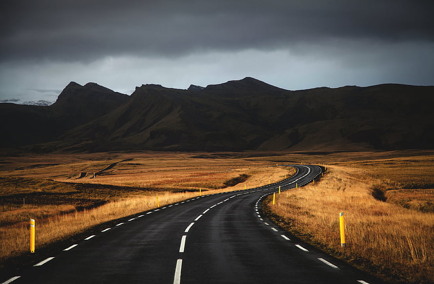 Winding asphalt road through golden grasslands under dramatic storm clouds and dark mountains HD wallpaper