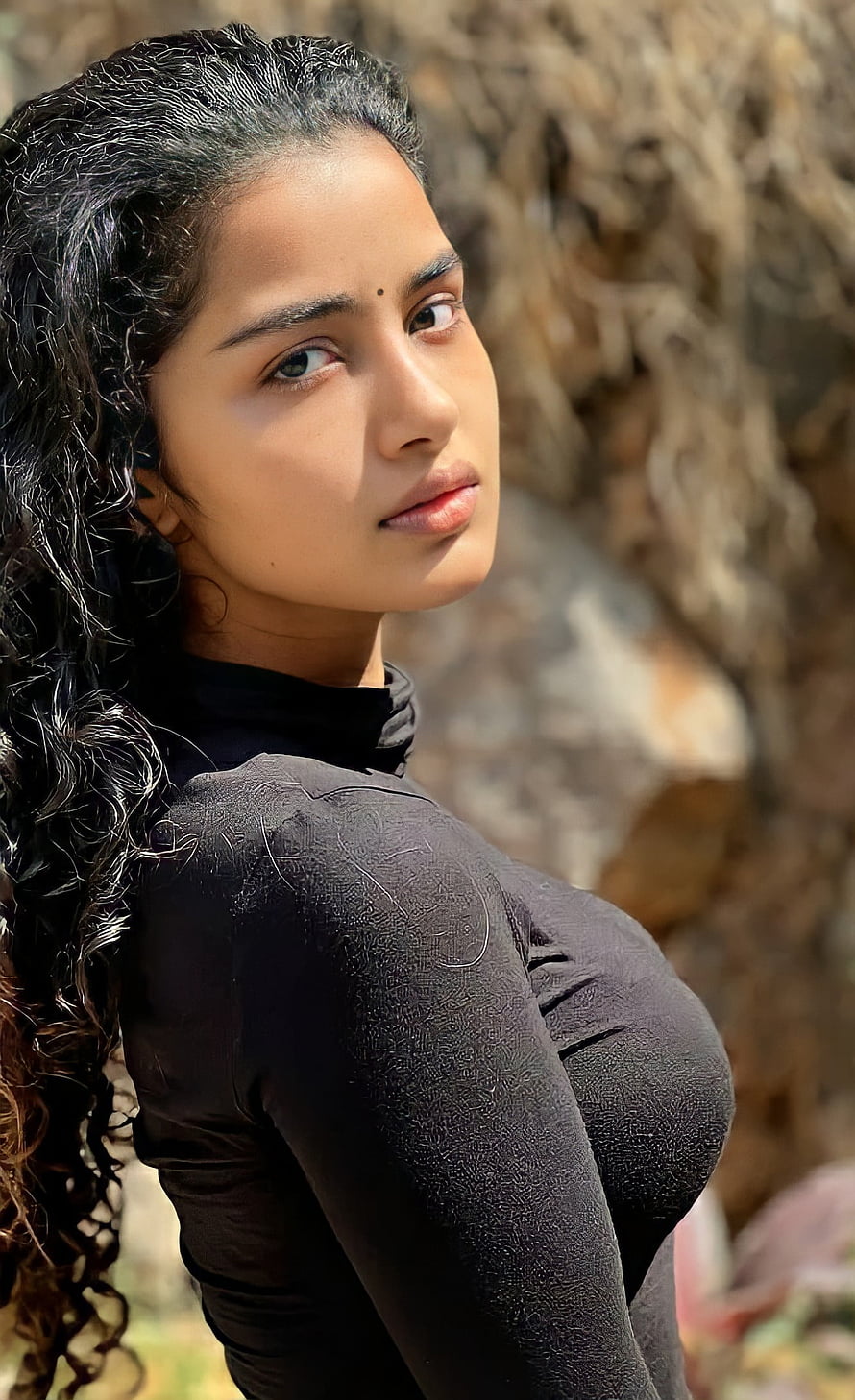 Anupama Parameswaran in a black turtleneck with long curly hair against a rocky background HD phone wallpaper