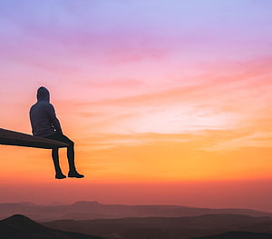 Silhouette of a person in a hoodie sitting on a wooden ledge overlooking a mountain range at sunset HD wallpaper