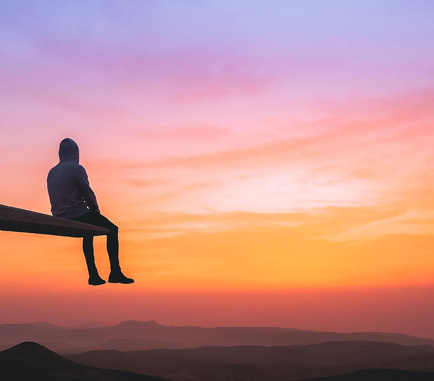 Silhouette of a person in a hoodie sitting on a wooden ledge overlooking a mountain range at sunset HD wallpaper