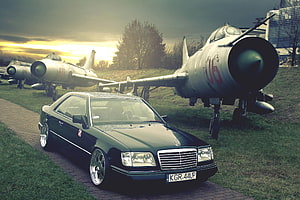 Black Mercedes-Benz W124 Coupe Parked in Front of Vintage Military Jets at Sunset HD wallpaper