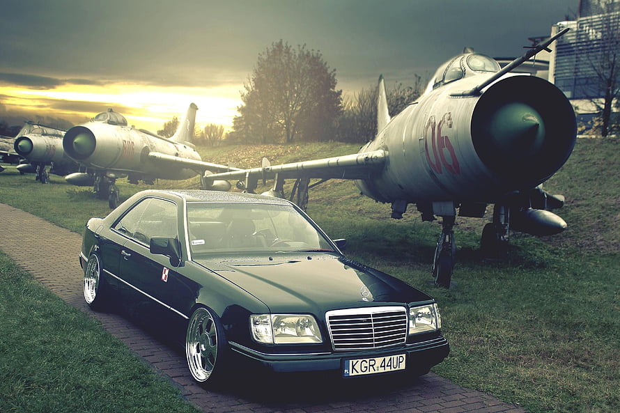 Black Mercedes-Benz W124 Coupe Parked in Front of Vintage Military Jets at Sunset HD wallpaper