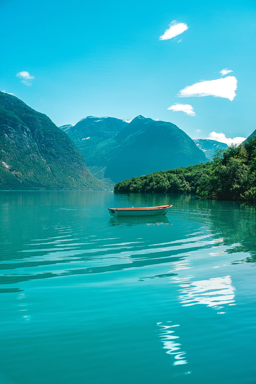 Serene turquoise lake with a small wooden boat floating near lush green mountains under a bright blue sky HD phone wallpaper