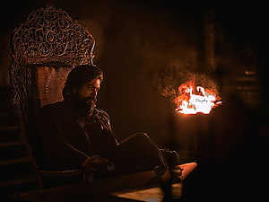 Yash in KGF seated on ornate throne beside a burning paper in dim cinematic lighting HD wallpaper