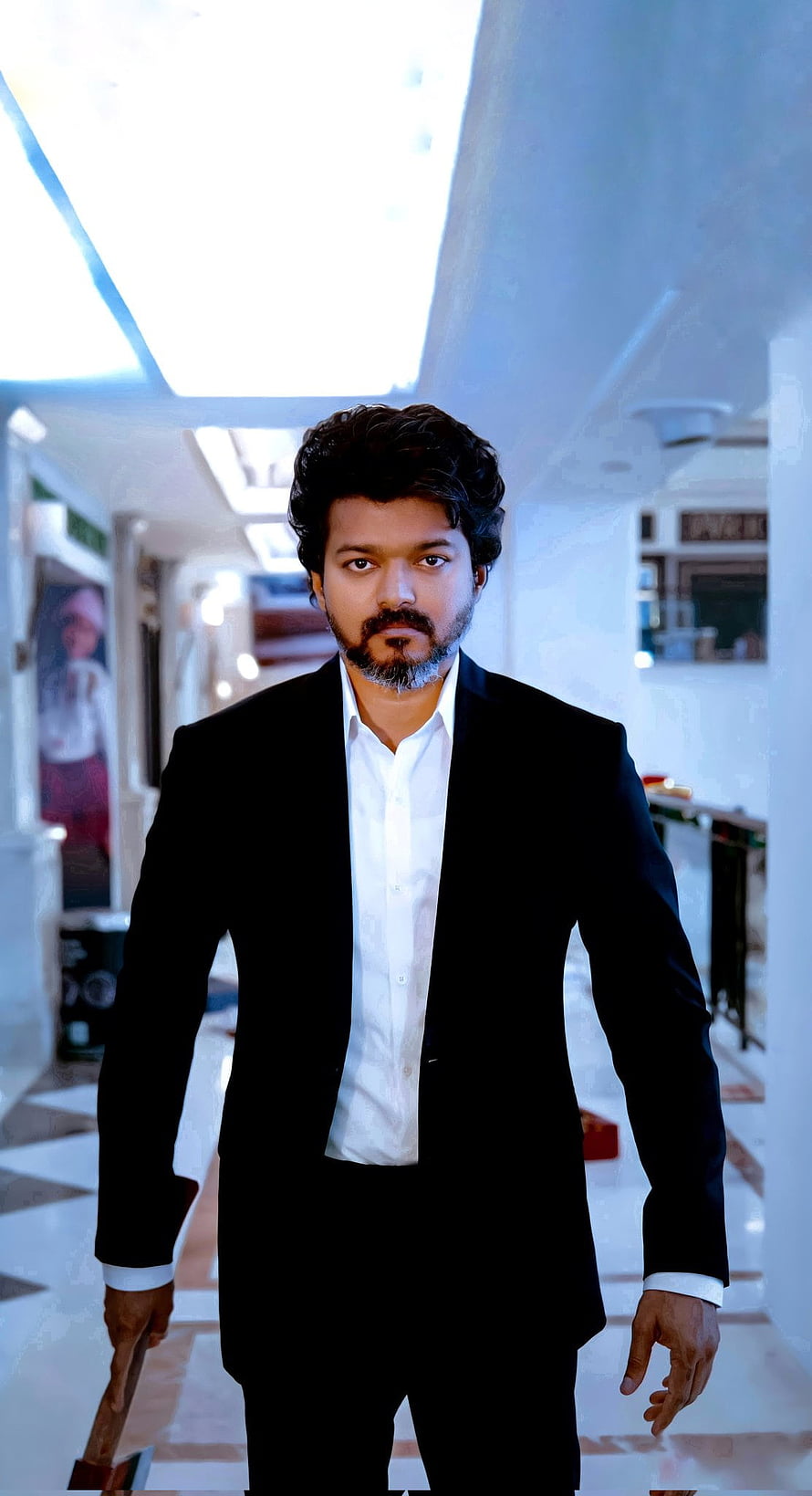 Thalapathy Vijay in a black suit and white shirt walking through a bright indoor corridor HD phone wallpaper