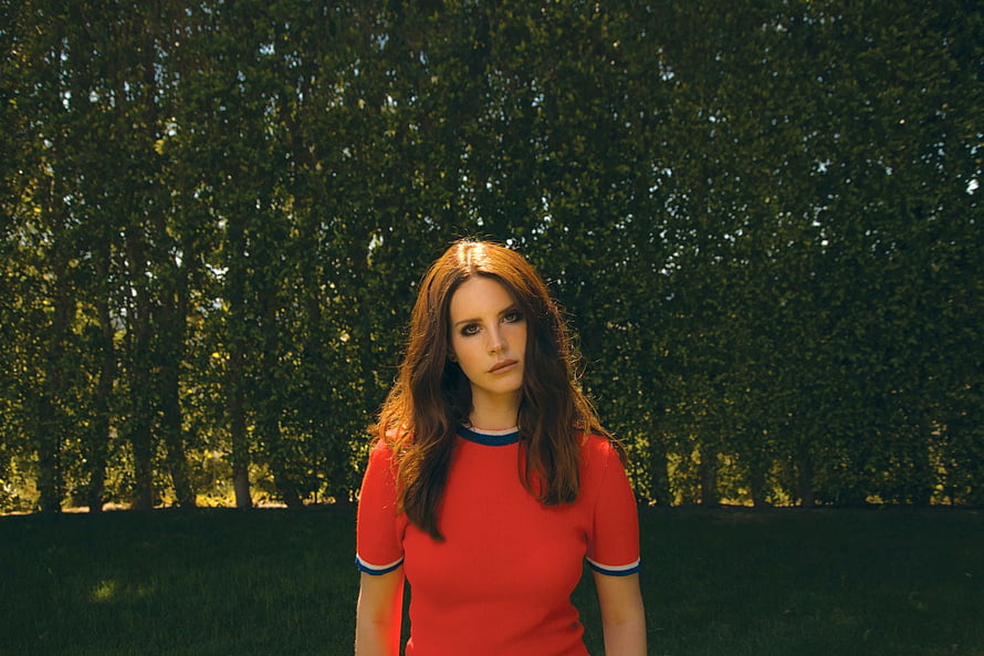 Lana Del Rey in a red dress standing in a sunlit park with a backdrop of tall green trees HD wallpaper
