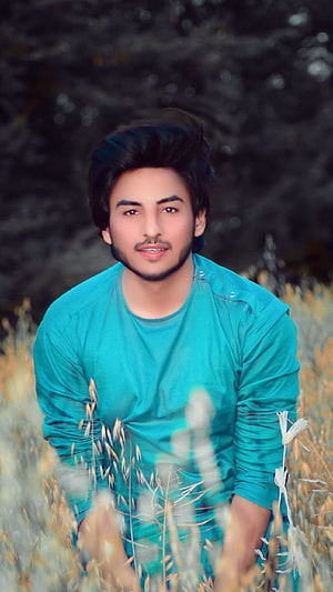 Young man in teal shirt sitting in a field of dry grass with dark foliage background HD phone wallpaper