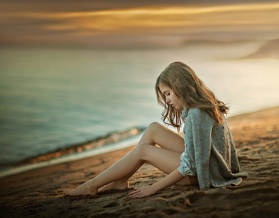 Young girl sitting on a sandy beach at sunset with a calm ocean backdrop HD wallpaper