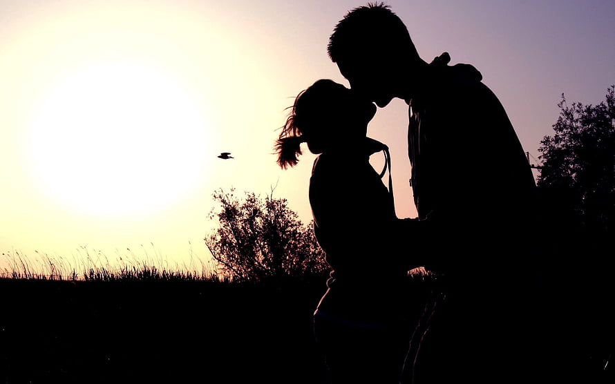 Silhouetted couple sharing a kiss against a golden sunset sky in a romantic field HD wallpaper