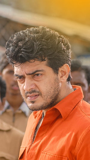 Ajith Kumar in an orange shirt with a serious expression and curly hair HD phone wallpaper