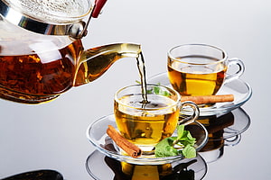 Glass teapot pouring amber tea into clear cups with cinnamon sticks and fresh mint leaves HD wallpaper
