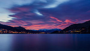 Vibrant Sunset Sky Over a Calm Lake with Distant Snow-Capped Mountains and Town Lights HD wallpaper