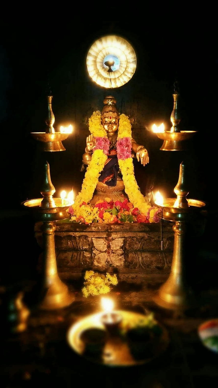 Ayyappan deity statue adorned with marigold garlands and lit brass oil lamps in a dark temple setting HD phone wallpaper