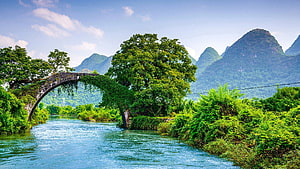 Ancient stone arch bridge over a calm river with lush karst mountains in the background HD wallpaper