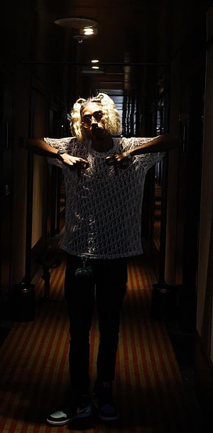 MC Stan in a white patterned shirt and glasses standing in a dimly lit hotel corridor HD phone wallpaper