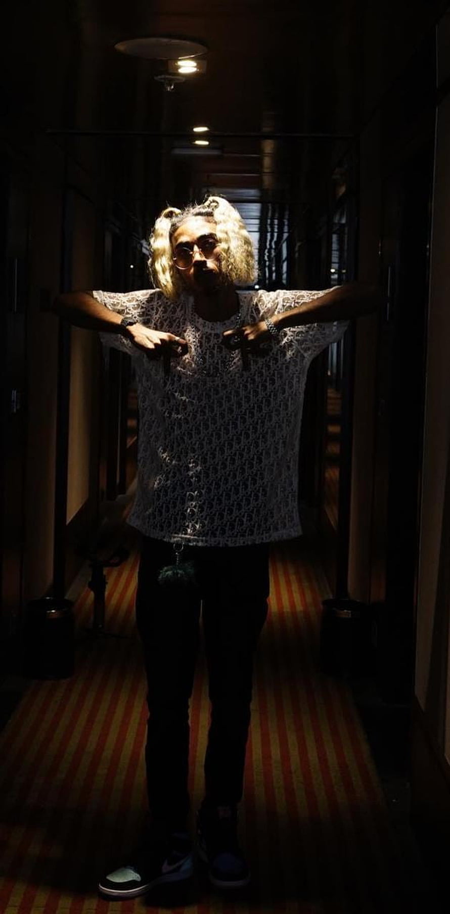 MC Stan in a white patterned shirt and glasses standing in a dimly lit hotel corridor HD phone wallpaper