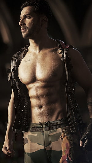 Varun Dhawan in a rugged open vest and camouflage pants with dramatic lighting HD phone wallpaper