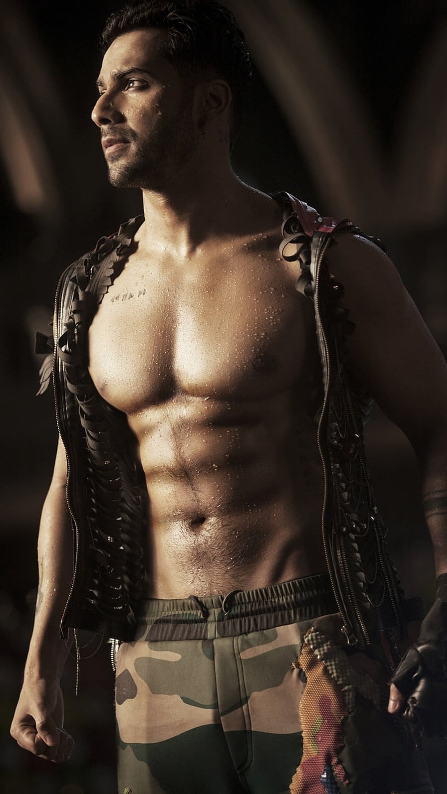 Varun Dhawan in a rugged open vest and camouflage pants with dramatic lighting HD phone wallpaper