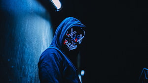 Anonymous figure in a dark hoodie wearing a glowing LED skull mask with red eyes in a moody blue-lit setting HD wallpaper