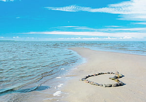 Heart-shaped stone arrangement on a serene sandy beach with gentle ocean waves and a bright blue sky HD wallpaper