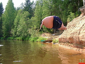 Man performing a backflip off a riverbank cliff into a forest lake HD wallpaper