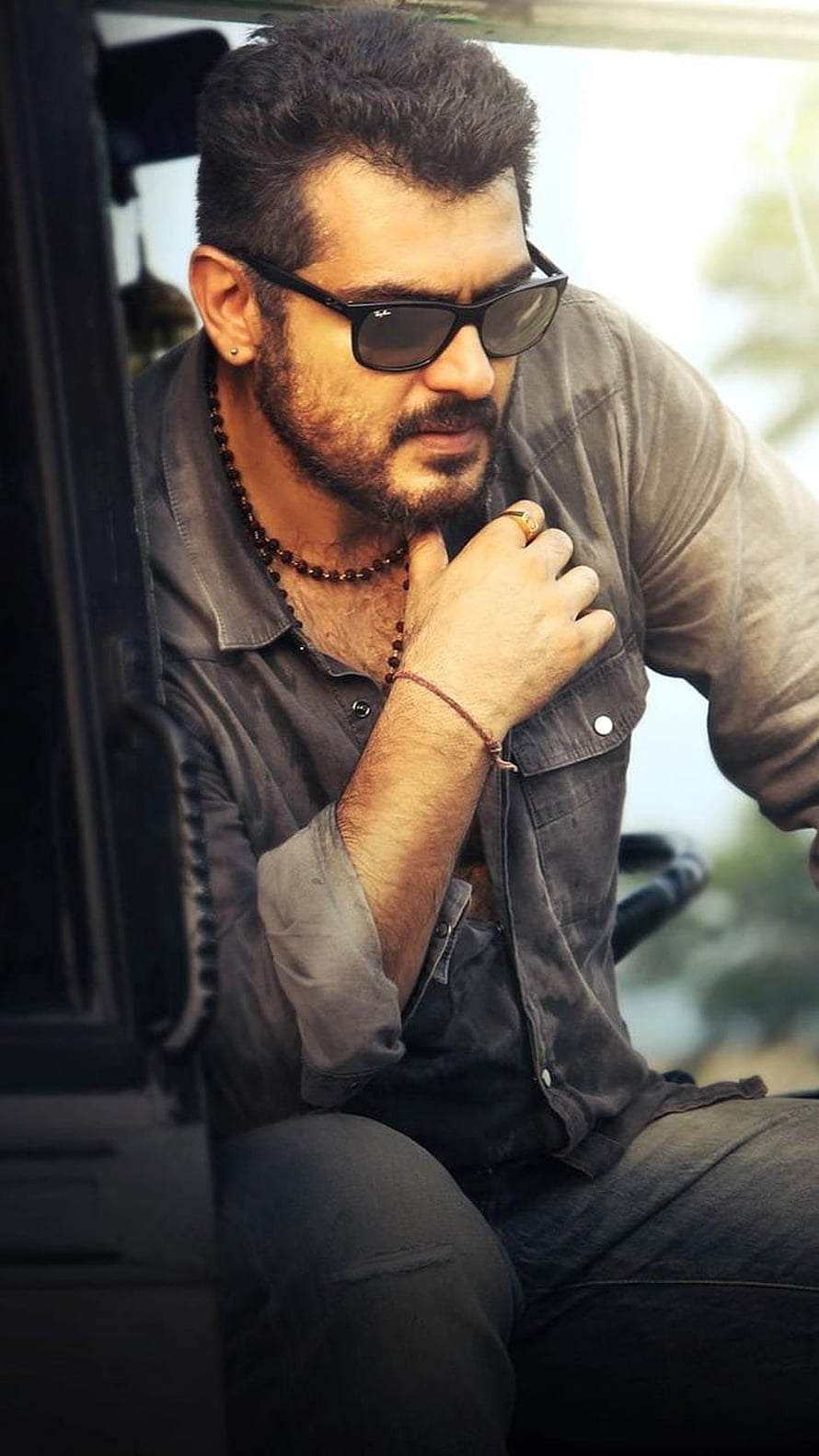 Ajith Kumar in a rugged grey shirt and sunglasses, seated in a vehicle with a contemplative expression HD phone wallpaper
