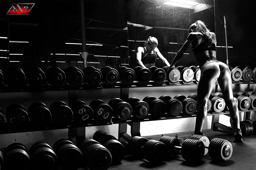 Black and white fitness model selecting dumbbells in a dark gym with mirror reflection HD wallpaper
