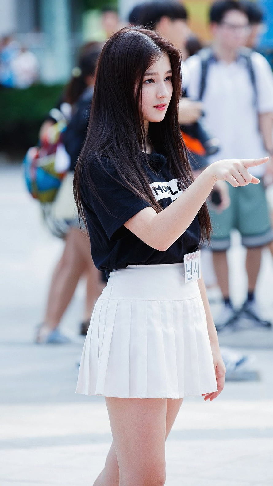 Nancy from MAMAMOO in a black t-shirt and white pleated skirt pointing outdoors HD phone wallpaper