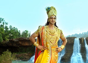 Krishna from Mahabharat in ornate golden attire standing before a scenic waterfall HD wallpaper