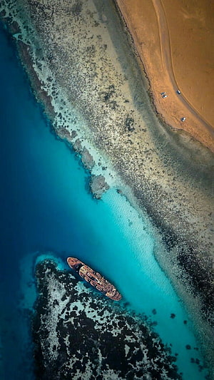 Aerial drone view of a shipwreck resting on a coral reef beside a sandy tropical beach HD phone wallpaper