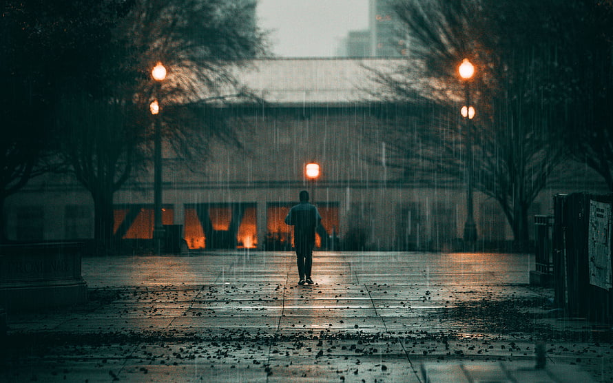 Solitary figure walking through a rainy city plaza at dusk with glowing street lamps HD wallpaper