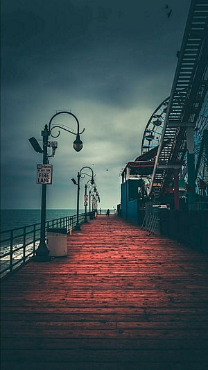 Moody wooden pier walkway with vintage street lamps and a Ferris wheel under a dark overcast sky HD phone wallpaper