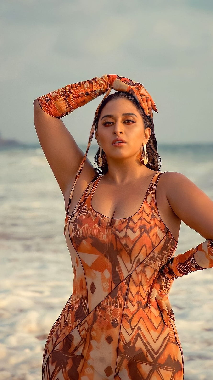 Raja Kumari in an orange patterned dress posing on a beach at sunset HD phone wallpaper