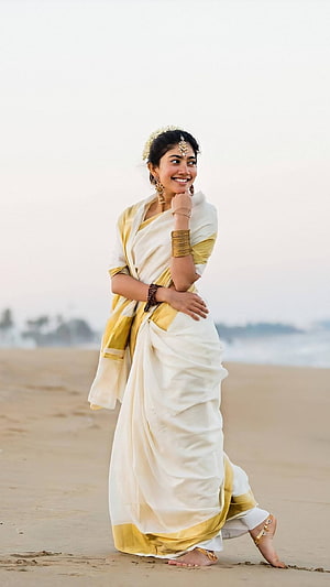 Sai Pallavi in a white and gold saree walking along a sandy beach HD phone wallpaper