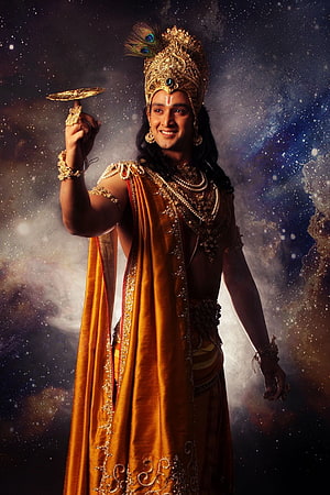 Lord Krishna in ornate golden crown and saffron robes holding a discus against a cosmic starry backdrop HD phone wallpaper