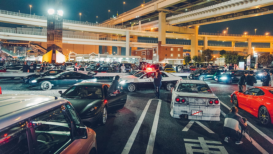 Nighttime car meet at Daikoku PA with classic and modern sports cars under highway overpass HD wallpaper