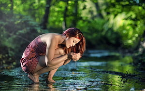 Red-haired woman crouching in a forest stream, cupping water in her hands HD wallpaper