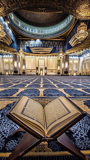Open Quran on a wooden stand inside a grand mosque with ornate golden dome and patterned carpet HD phone wallpaper